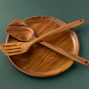 A handcrafted teak wood kitchen set featuring a round wooden plate 30 cm, a solid medium spatula, and a slotted fish slice turner crossed on top. The set is isolated against a matte dark green background, highlighting the rich, matching golden-brown grain patterns of the premium teak. This sustainable and non-scratch utensil collection is designed for versatile cooking and elegant serving in a modern, eco-friendly kitchen.
