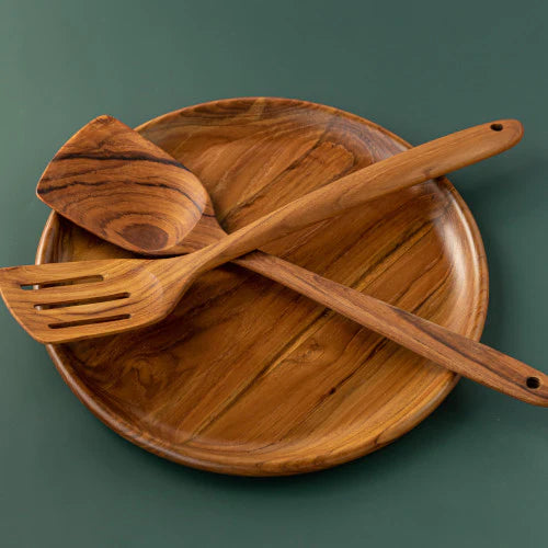 A handcrafted teak wood kitchen set featuring a round wooden plate 30 cm, a solid medium spatula, and a slotted fish slice turner crossed on top. The set is isolated against a matte dark green background, highlighting the rich, matching golden-brown grain patterns of the premium teak. This sustainable and non-scratch utensil collection is designed for versatile cooking and elegant serving in a modern, eco-friendly kitchen.