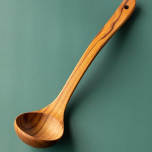 A professional product photograph of a handcrafted large teak wood ladle, positioned diagonally across a smooth, solid sage-green background. The utensil features a deep, rounded bowl and a long, gently curved ergonomic handle with a functional hanging hole at the tip. The shot beautifully showcases the warm golden-brown timber tones and prominent, wavy natural grain patterns of the single-piece, sustainable teak construction.