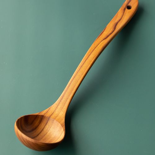 A professional product photograph of a handcrafted large teak wood ladle, positioned diagonally across a smooth, solid sage-green background. The utensil features a deep, rounded bowl and a long, gently curved ergonomic handle with a functional hanging hole at the tip. The shot beautifully showcases the warm golden-brown timber tones and prominent, wavy natural grain patterns of the single-piece, sustainable teak construction.