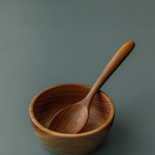A top-down lifestyle photograph of two handcrafted teak wood bowls of different sizes on a dark, textured slate-grey surface. The larger bowl is filled with whole coriander seeds and holds a small wooden scoop, while a smaller companion bowl contains bright red chili powder with its own wooden spoon. The image highlights the warm, natural grain of the sustainable teak, showcasing the bowls' versatility as both a durable toddler mealtime set and as artisanal kitchen tools for food preparation.