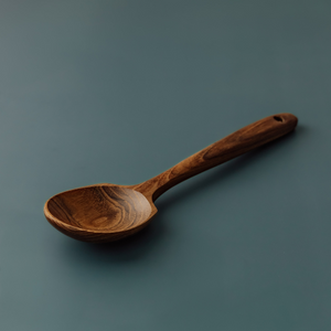 A close-up side-angle photograph of "My First Big Spoon," a handcrafted teak wood spoon for children, resting on a smooth, muted grey-blue surface. The image highlights the spoon's signature wide, shallow head and thick, ergonomic handle, designed specifically for small hands to grip easily. The rich, warm natural grain of the sustainable teak wood is clearly visible, showcasing a smooth, non-toxic, and food-safe finish that is gentle on little mouths.