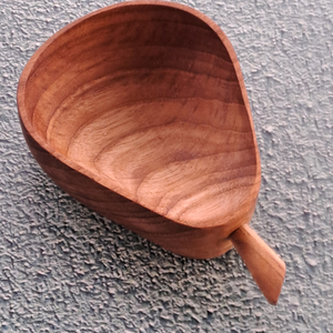 A top-down product photograph of a handcrafted pear-shaped dip bowl carved from sustainable teak wood, angled diagonally against a neutral, textured grey wall surface. The 10cm long dish features a wide rounded base tapering into a narrower neck with a small, stylized wooden stem handle at the tip. The image captures the deep, smooth interior basin, highlighting the rich golden-brown timber tones and prominent, rippled natural wood grain patterns of the non-toxic, food-safe single-piece mini bowl.