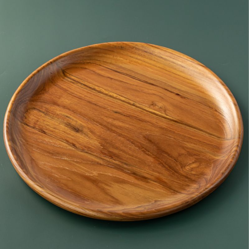 A handcrafted, round teak wood plate or serving platter, 25 cm in diameter isolated on a smooth, dark forest green background. The image showcases the rich, swirling natural wood grain with deep golden-brown and amber tones, highlighting the plate's polished finish and organic, slightly irregular rim. This durable, sustainable wooden dinnerware is ideal for serving appetizers or side dishes.
