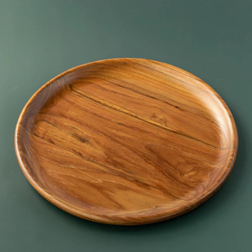 A handcrafted, round teak wood plate or serving platter, 30 cm in diameter isolated on a smooth, dark forest green background. The image showcases the rich, swirling natural wood grain with deep golden-brown and amber tones, highlighting the plate's polished finish and organic, slightly irregular rim. This durable, sustainable wooden dinnerware is ideal for serving appetizers or side dishes.