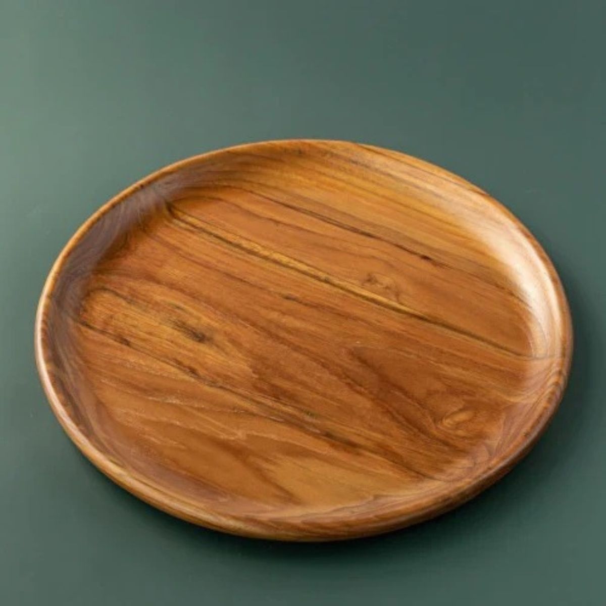 A handcrafted, round teak wood plate or serving platter, 35 cm in diameter isolated on a smooth, dark forest green background. The image showcases the rich, swirling natural wood grain with deep golden-brown and amber tones, highlighting the plate's polished finish and organic, slightly irregular rim. This durable, sustainable wooden dinnerware is ideal for serving appetizers or side dishes.
