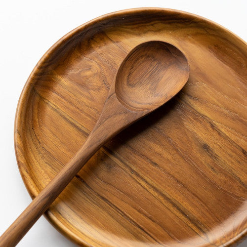An overhead close-up photograph of a handcrafted long-handled teak wood spoon resting across a matching large teak wood platter. Both pieces feature a vibrant, swirling natural grain in warm golden and chestnut tones, set against a deep forest green background. Carved from solid, sustainable teak, this duo highlights a coordinated, food-safe, and non-toxic aesthetic for serving large meals or appetizers in a sustainable kitchen.