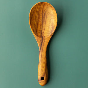 A top-down product photograph of a handcrafted 22cm teak wood rice paddle and grain serving spoon, centered vertically against a smooth, matte green surface. The utensil features a wide, shallow bowl and a short, ergonomically thick handle with a functional hanging hole at the base. The image highlights the rich, swirling golden-brown natural grain of the single-piece sustainable teak, showcasing its smooth, non-stick design for fluffing and serving rice or grains.