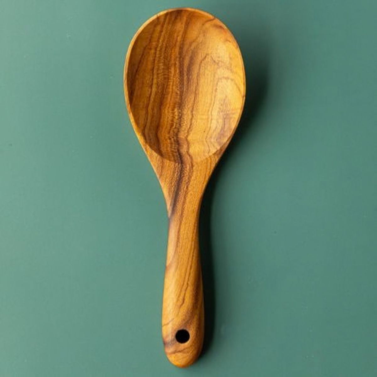 A top-down product photograph of a handcrafted 22cm teak wood rice paddle and grain serving spoon, centered vertically against a smooth, matte green surface. The utensil features a wide, shallow bowl and a short, ergonomically thick handle with a functional hanging hole at the base. The image highlights the rich, swirling golden-brown natural grain of the single-piece sustainable teak, showcasing its smooth, non-stick design for fluffing and serving rice or grains.