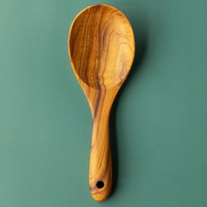 A top-down product photograph of a handcrafted 22cm teak wood rice paddle and grain serving spoon, centered vertically against a smooth, matte green surface. The utensil features a wide, shallow bowl and a short, ergonomically thick handle with a functional hanging hole at the base. The image highlights the rich, swirling golden-brown natural grain of the single-piece sustainable teak, showcasing its smooth, non-stick design for fluffing and serving rice or grains.