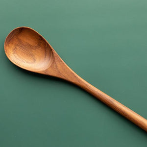 A top-down product photograph of a handcrafted teak wood rice serving spoon and paddle, centered vertically against a smooth, solid sage-green background. The utensil features a wide, gently hollowed oval bowl and a short, thick ergonomic handle with a clean hanging hole at the base. The composition emphasizes the rich, contrasting golden-brown tones and swirling natural timber grain unique to this single-piece, sustainable teak tool.