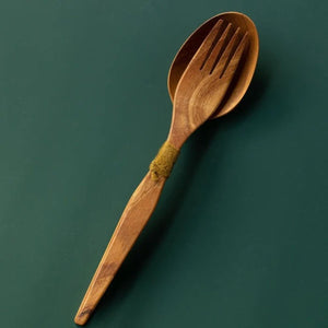 Handcrafted teak wood salad server set with a serving fork nested inside a spoon tied with olive green yarn, shown from a top-down view on a dark green surface.