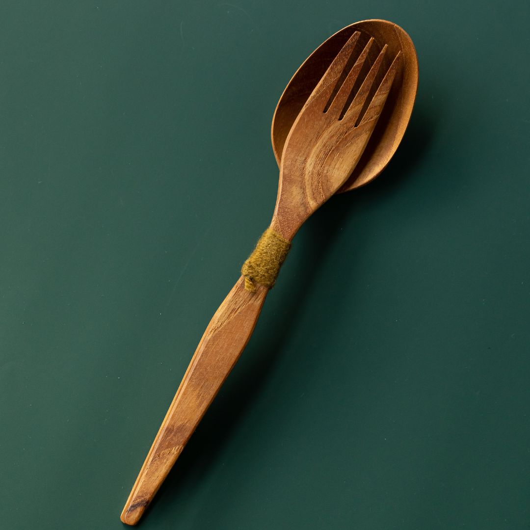 Handcrafted teak wood salad server set with a serving fork nested inside a spoon tied with olive green yarn, shown from a top-down view on a dark green surface.