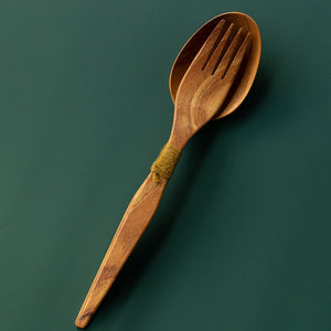 Handcrafted teak wood salad server set with a serving fork nested inside a spoon tied with olive green yarn, shown from a top-down view on a dark green surface.