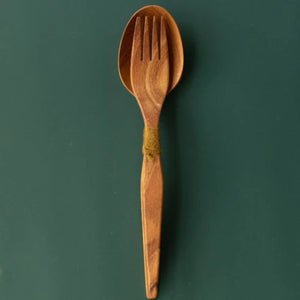 Handcrafted teak wood salad server set with a fork nested in a spoon tied with green yarn, shown from a top-down view on a dark green surface.
