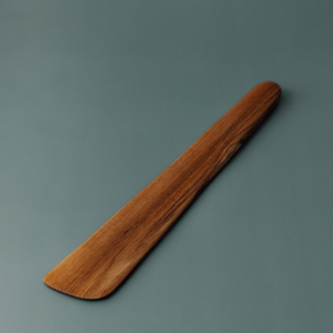 A handcrafted teak wood sauté spatula and scraper featuring a wide, flat-edged blade designed for effortless stirring, flipping, and deglazing. The spatula is carved from a single piece of durable, golden-brown teak wood with a rich natural grain and a smooth, food-safe finish. This long-handled, non-toxic kitchen tool is ideal for use with non-stick cookware. stainless steel, cast iron and provides an ergonomic grip for everyday cooking in a sustainable, artisanal kitchen.