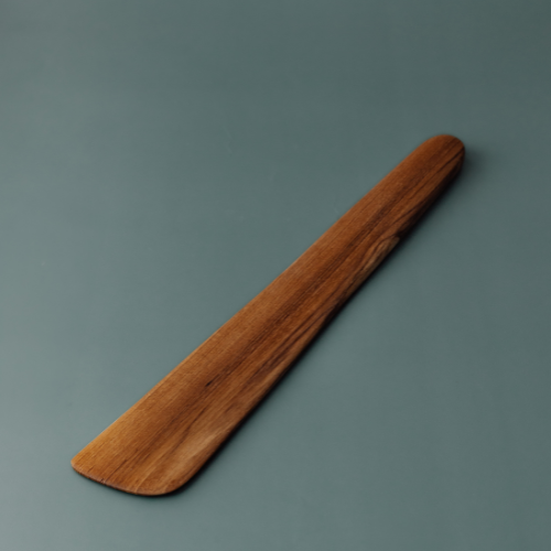 A handcrafted teak wood sauté spatula and scraper featuring a wide, flat-edged blade designed for effortless stirring, flipping, and deglazing. The spatula is carved from a single piece of durable, golden-brown teak wood with a rich natural grain and a smooth, food-safe finish. This long-handled, non-toxic kitchen tool is ideal for use with non-stick cookware. stainless steel, cast iron and provides an ergonomic grip for everyday cooking in a sustainable, artisanal kitchen.