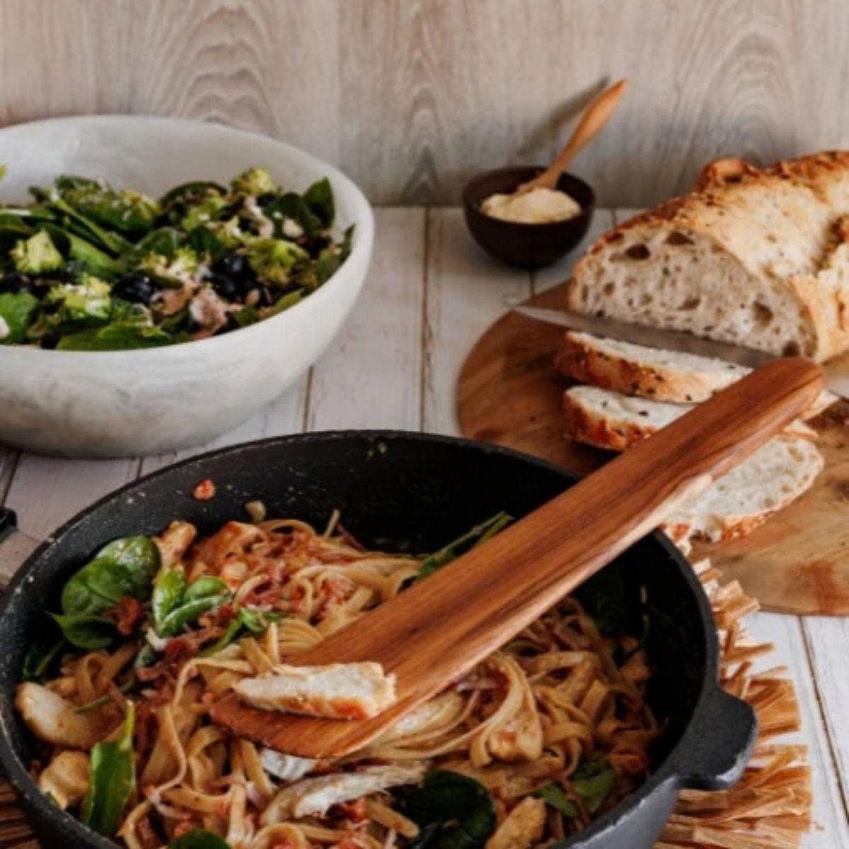 A close-up culinary lifestyle photograph of a 37cm handcrafted teak wood sauté spatula lifting a serving of chicken and spinach pasta from a black skillet. Strands of fettuccine, fresh spinach leaves, and chicken are balanced on the flat blade of the spatula. In the soft-focus background, a rustic table setting features a fresh green salad in a large bowl and sliced sourdough bread. The image showcases the rich natural grain of the single-piece teak tool, emphasizing its food-safe, non-toxic, and functional