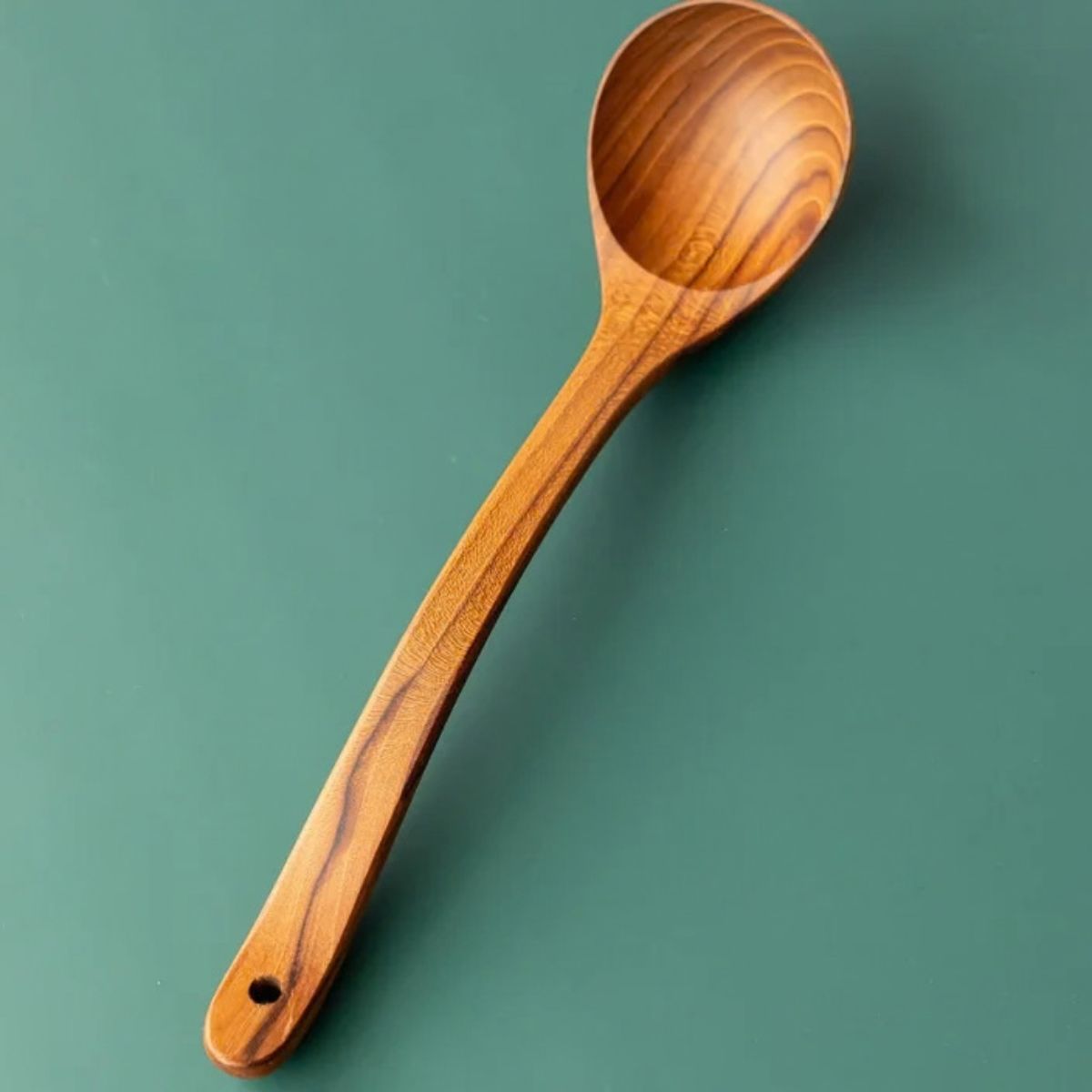 Top-down view of a handcrafted teak wood serving ladle with a long ergonomic handle and deep round bowl, featuring a natural wood grain finish on a dark green surface.