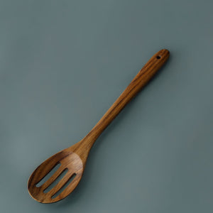 A full-length product photograph of a handcrafted slotted serving spoon, positioned diagonally across a smooth, solid teak background. Carved from a single piece of sustainable teak wood, the utensil features a slotted oval head with three precision drainage slots and a long, smooth ergonomic handle complete with a functional hanging hole near the tip. The studio lighting highlights the rich golden-brown timber tones and flowing natural wood grain of this non-toxic, food-safe kitchen tool.
