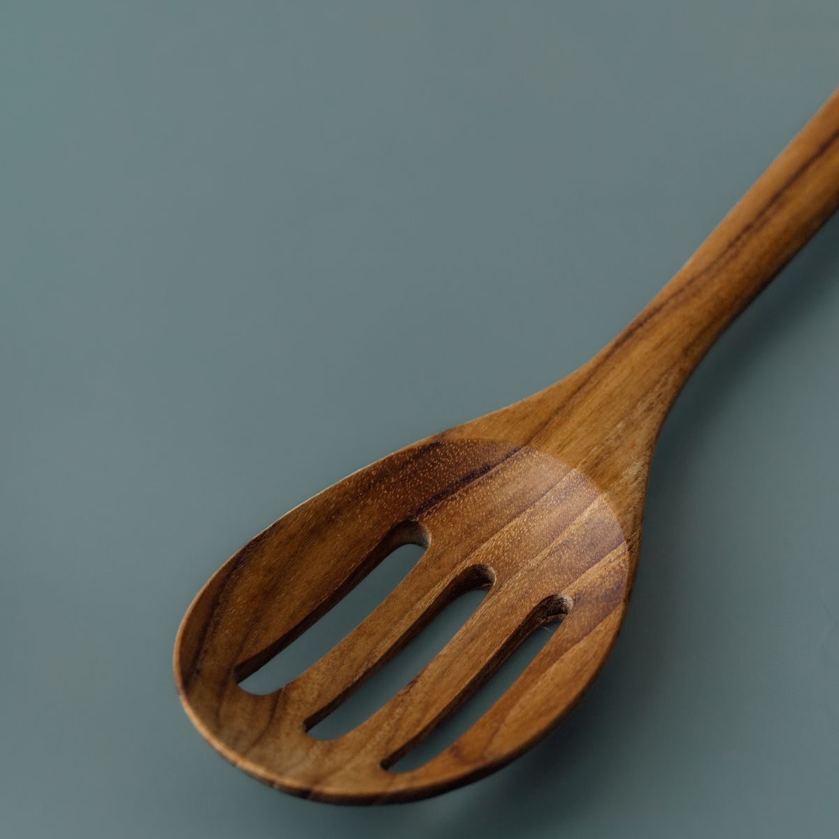 A close-up macro photograph focusing on the head of a handcrafted slotted serving spoon carved from sustainable teak wood, angled against a smooth, solid slate-grey background. The image highlights the detailed craftsmanship of the three smooth drainage slots, the shallow curved scoop, and the soft bevel along the outer rim. Soft studio lighting showcases the rich, warm golden-brown timber tones and fine, smooth finish of the non-toxic, single-piece wooden utensil.