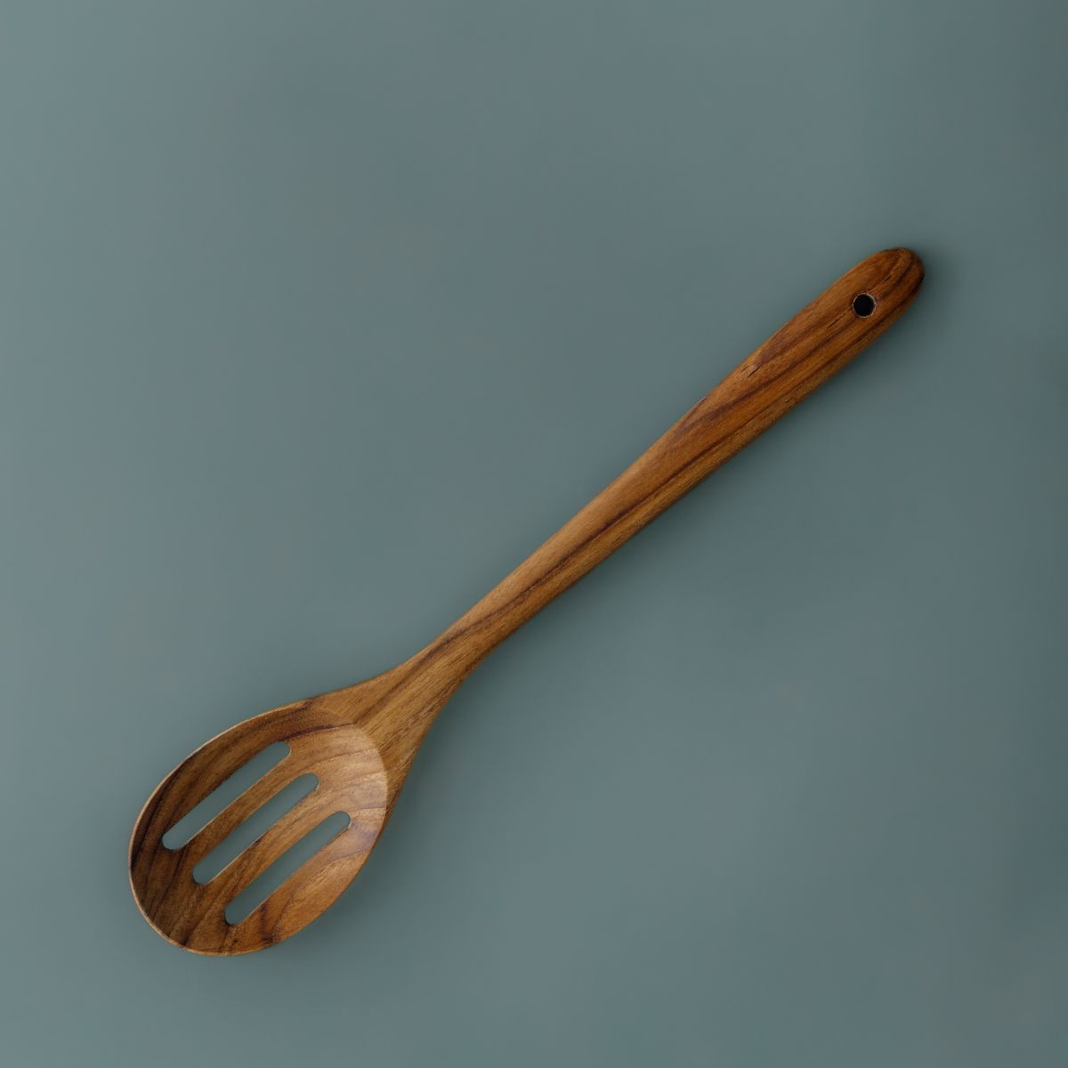 A full-length product photograph of a handcrafted slotted serving spoon angled centrally against a smooth, solid slate-grey studio backdrop. Carved from a single piece of sustainable teak wood, the utensil features a wide slotted head with three smooth drainage slots, leading up a long, contoured handle with a functional hanging hole at the top. The overhead lighting showcases the rich golden-brown hues and deep natural grain variations of this non-toxic, food-safe wooden utensil.