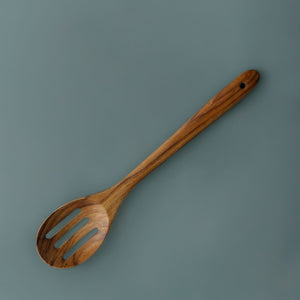 A full-length product photograph of a handcrafted slotted serving spoon angled centrally against a smooth, solid slate-grey studio backdrop. Carved from a single piece of sustainable teak wood, the utensil features a wide slotted head with three smooth drainage slots, leading up a long, contoured handle with a functional hanging hole at the top. The overhead lighting showcases the rich golden-brown hues and deep natural grain variations of this non-toxic, food-safe wooden utensil.