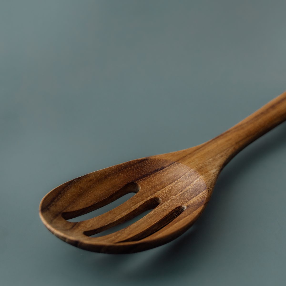 A close-up product photograph of a handcrafted slotted serving spoon carved from sustainable teak wood, positioned diagonally against a smooth, solid slate-grey background. The image focuses on the head of the utensil, featuring a wide, gently curved oval bowl with three smooth drainage slots and a slim handle. The studio lighting showcases the rich golden-brown timber tones, subtle grain variation, and smooth, food-safe finish of the single-piece wooden tool