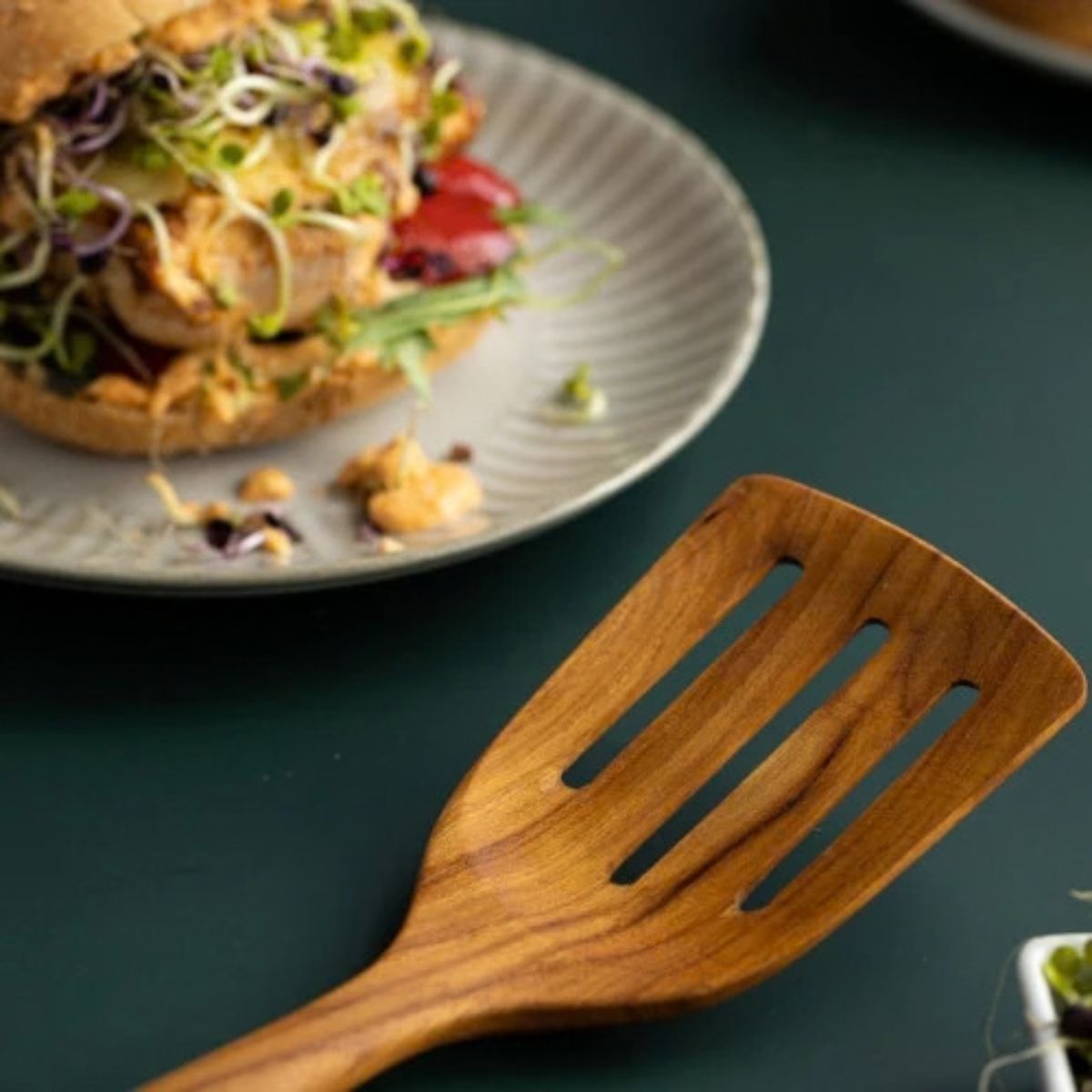 A detailed close-up shot of the wide head of a handcrafted teak wood slotted turner, resting on a matte teal surface. The image clearly shows the three precise drainage slots and highlights the rich, natural wood grain. In the softly blurred background, a gourmet veggie burger on a grey plate illustrates a practical use for this durable, non-scratch kitchen utensil, which is designed for flipping and serving without damaging cookware.