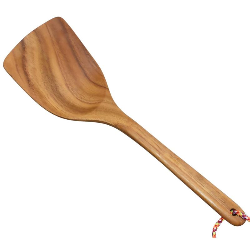 A handcrafted small teak wood spatula, 23 cm in length, featuring a wide, angled head for precise flipping and stirring. The spatula is carved from a single piece of golden-brown teak wood with a visible natural grain and a smooth, food-safe finish. It includes a slim, ergonomic handle with a colorful hanging cord at the end for easy storage. This compact, non-toxic kitchen tool is ideally sized for small pans, eggs, or serving desserts in a sustainable kitchen.