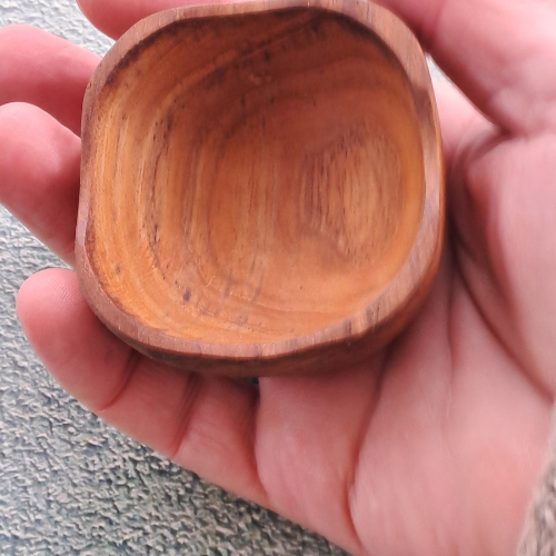 A close-up lifestyle photograph showing a small, square handcrafted pinch bowl carved from sustainable teak wood resting in the open palm of a person's hand. The overhead perspective clearly demonstrates the compact size and scale of the dish, highlighting its softly rounded square rim, deep smooth interior, and rich golden-brown timber tones with a prominent natural wood grain. Carved from a single piece of non-toxic teak, this durable mini pot is designed for holding spices, salts, or condiments.