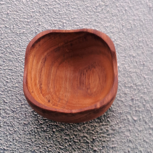 A top-down product photograph of a small, square handcrafted pinch bowl carved from sustainable teak wood, centered against a neutral, textured grey wall surface. The image highlights the unique, softly rounded square rim and deep, smooth interior of the dish, showcasing the rich golden-brown timber tones and prominent natural wood grain patterns. Expertly shaped from a single piece of non-toxic, food-safe teak, this durable mini bowl is designed as a functional culinary essential for holding spices, salts,