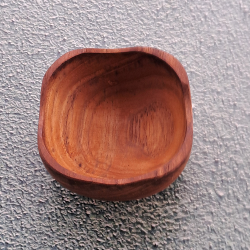 A top-down product photograph of a small, square handcrafted pinch bowl carved from sustainable teak wood, centered against a neutral, textured grey wall surface. The image highlights the unique, softly rounded square rim and deep, smooth interior of the dish, showcasing the rich golden-brown timber tones and prominent natural wood grain patterns. Expertly shaped from a single piece of non-toxic, food-safe teak, this durable mini bowl is designed as a functional culinary essential for holding spices, salts,