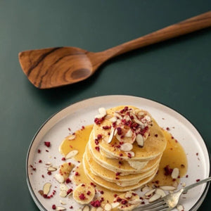 A lifestyle shot featuring a handcrafted teak wood spatula resting on a dark green surface next to a stack of fresh pancakes topped with sliced bananas, almonds, and syrup. The image highlights the spatula's beautiful dark wood grain and wide turner head, demonstrating its use as a gentle, non-scratch tool perfect for flipping delicate breakfast items like pancakes, crepes, and eggs.