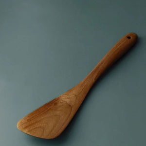 A detailed close-up shot of a handcrafted teak wood spatula head resting on a smooth, matte teal surface. The image highlights the wide, flat turner head and the rich, natural wood grain patterns of the premium teak. This perspective emphasizes the smooth, polished finish and the sturdy construction of the utensil, showcasing it as a high-quality, non-scratch tool for flipping and serving in a modern kitchen.