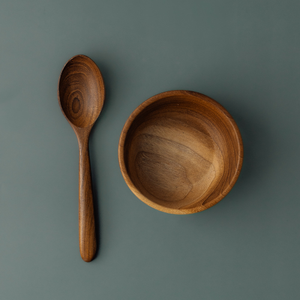 A top-down product photograph of a handcrafted 11 cm teak wood toddler bowl paired with a matching rounded wooden spoon. The items are laid out symmetrically on a smooth, muted grey-blue surface, highlighting the consistent, swirling natural grain of the sustainable teak. Both pieces are carved from single pieces of wood, featuring a non-toxic, food-safe finish. This durable and plastic-free set is designed for small hands, promoting independent eating in a healthy home environment.
