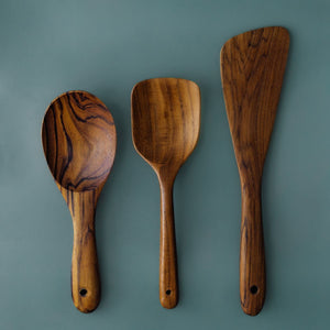 A full-length product photograph displaying a set of three handcrafted teak wood cooking utensils arranged vertically side-by-side against a smooth, solid slate-grey background. From left to right: a wide oval cooking spoon with prominent dark grain, a medium spatula with a rounded square head, and a long asymmetric sauté scraper spatula. Each utensil features a smooth ergonomic handle complete with a functional hanging hole at the base, showcasing the rich golden-brown timber and organic grain variations o