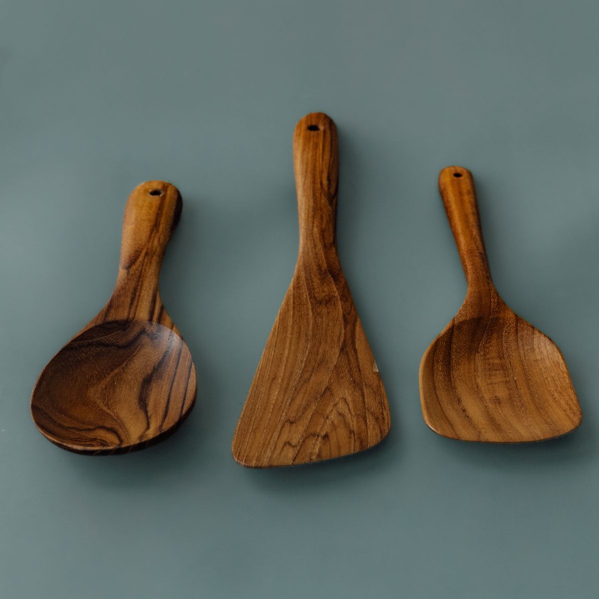 A full-length product photograph displaying a trio of handcrafted teak wood cooking utensils arranged side-by-side with handles pointing upwards against a smooth, solid slate-grey background. From left to right: a wide oval cooking spoon, a flat asymmetric sauté scraper spatula, and a rounded square spatula. The image showcases the smooth ergonomic handles with functional hanging holes at the top and emphasizes the rich, warm golden-brown timber tones and varied natural wood grain across each single-piece, 