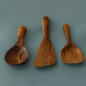 A full-length product photograph displaying a trio of handcrafted teak wood cooking utensils arranged side-by-side with handles pointing upwards against a smooth, solid slate-grey background. From left to right: a wide oval cooking spoon, a flat asymmetric sauté scraper spatula, and a rounded square spatula. The image showcases the smooth ergonomic handles with functional hanging holes at the top and emphasizes the rich, warm golden-brown timber tones and varied natural wood grain across each single-piece, 