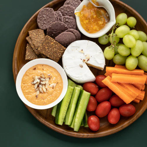 A top-down lifestyle photograph of a handcrafted teak wooden plate that is 20 cm's in diameter, used as a grazing platter on a dark green surface. The round platter is artfully arranged with a wedge of brie cheese, green grapes, baby carrots, cucumbers, cherry tomatoes,  crackers, and a small white bowl of hummus topped with almonds. The image highlights the plate's versatility as a premium, food-safe serving dish that complements vibrant, fresh ingredients in a modern kitchen or dining setting.