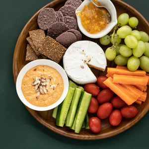 A top-down lifestyle photograph of a handcrafted teak wooden plate that is 30 cm's in diameter, used as a grazing platter on a dark green surface. The round platter is artfully arranged with a wedge of brie cheese, green grapes, baby carrots, cucumbers, cherry tomatoes,  crackers, and a small white bowl of hummus topped with almonds. The image highlights the plate's versatility as a premium, food-safe serving dish that complements vibrant, fresh ingredients in a modern kitchen or dining setting.