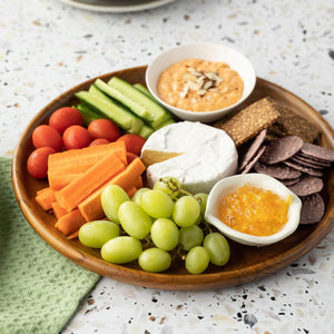 A top-down lifestyle photograph of a handcrafted teak wooden plate that is 25 cm's in diameter, used as a grazing platter on a dark green surface. The round platter is artfully arranged with a wedge of brie cheese, green grapes, baby carrots, cucumbers, cherry tomatoes,  crackers, and a small white bowl of hummus topped with almonds. The image highlights the plate's versatility as a premium, food-safe serving dish that complements vibrant, fresh ingredients in a modern kitchen or dining setting.