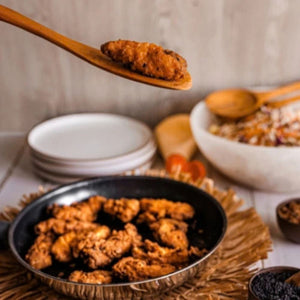 A lifestyle action shot of a handcrafted wooden cooking spoon lifting a piece of golden-brown fried chicken from a black non-stick frying pan. The background features a rustic kitchen setting with a white salad bowl, a stack of ceramic plates, and small bowls of spices on a wooden tabletop. This image demonstrates the heat-resistant and non-scratch properties of teak wood utensils, making them ideal for frying, sautéing, and serving hot meals.