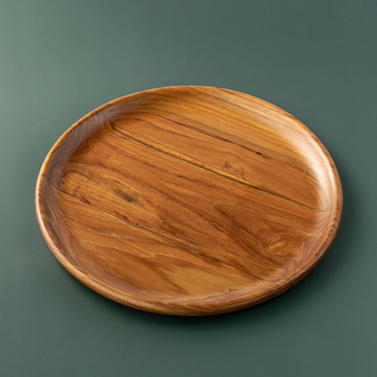 A handcrafted, round teak wood plate or serving platter, 15 cm in diameter isolated on a smooth, dark forest green background. The image showcases the rich, swirling natural wood grain with deep golden-brown and amber tones, highlighting the plate's polished finish and organic, slightly irregular rim. This durable, sustainable wooden dinnerware is ideal for serving appetizers or side dishes.
