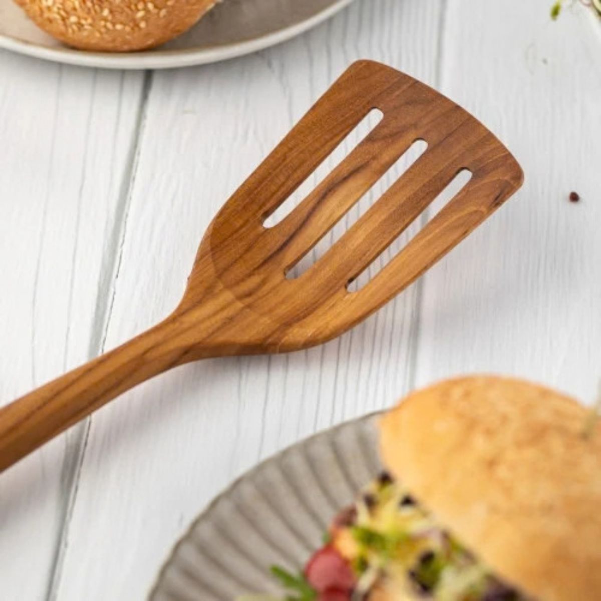 A close-up lifestyle shot of a handcrafted teak wood slotted turner resting on a white wooden table next to a fresh gourmet burger on a ceramic plate. The image highlights the wide, three-slotted head and the rich natural wood grain of the turner, emphasizing its use as a durable, non-scratch tool for flipping and serving burgers, eggs, and delicate proteins. The bright, clean kitchen aesthetic showcases the product as both a functional cooking utensil and an artisanal serving piece