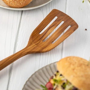 A close-up lifestyle shot of a handcrafted teak wood slotted turner resting on a white wooden table next to a fresh gourmet burger on a ceramic plate. The image highlights the wide, three-slotted head and the rich natural wood grain of the turner, emphasizing its use as a durable, non-scratch tool for flipping and serving burgers, eggs, and delicate proteins. The bright, clean kitchen aesthetic showcases the product as both a functional cooking utensil and an artisanal serving piece