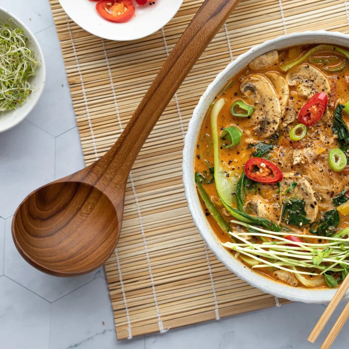 A bowl of prepared chicken and mushroom soup with vegetables, placed on a bamboo placemat beside a large wooden spoon, with small bowls of garnish in the background.
