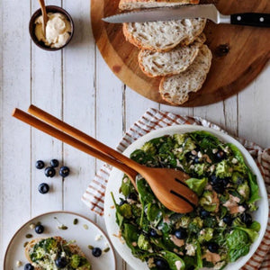 Handcrafted large wooden salad servers with long handles resting in a white ceramic bowl filled with a fresh green spinach and broccoli salad