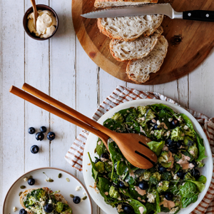 Handcrafted large wooden salad servers with long handles resting in a white ceramic bowl filled with a fresh green spinach and broccoli salad