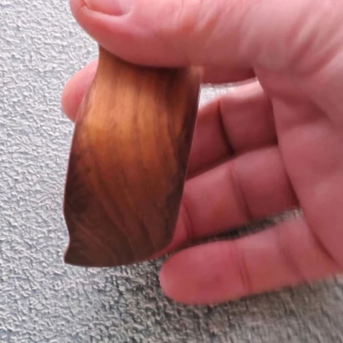 A close-up side perspective lifestyle photograph of a small, square handcrafted pinch bowl carved from sustainable teak wood, held between a person's thumb and fingers against a neutral textured grey background. The shot highlights the exterior profile of the dish, emphasizing its smooth, thick outer walls and gently curved vertical edge. The image features the rich, warm golden-brown timber finish and flowing natural wood grain patterns of the durable, single-piece mini bowl.